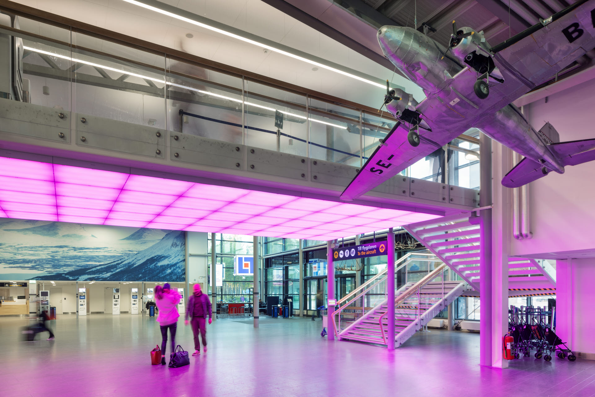 Terminalen på Luleå flygplats, tidigare kallad Kallax. En stor modell av en äldre flygplanstyp hänger i taket i avgångshallen. Det är glest mellan människor, en person håller i ett litet barn på håll.